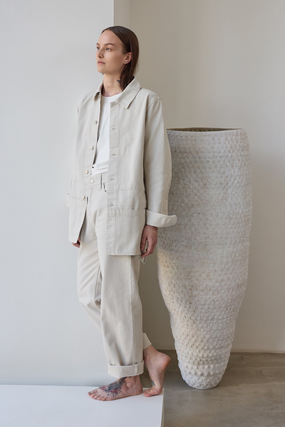 Nomen Nescio 320 Worker Denim Jacket white – model lifestyle, leaning against wall with ceramic sculpture